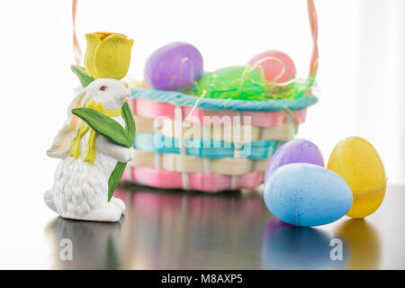 An Easter Bunny is set a long side his basket filled with Easter Eggs. Plastic eggs are also groupled in front of the basket as their reflections are  Stock Photo