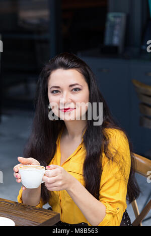 Female with long dark hair wearing yellow shirt and red lipstik in a cafe with dark interior, holding a cup of cappuccino in her hands. Stock Photo
