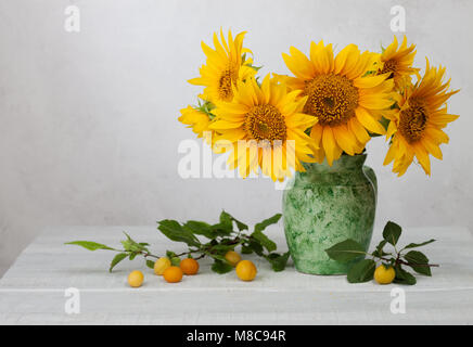 Bouquet of sunflowers in old ceramic jug against a white wooden wall. In the foreground branches with ripe cherry plum Stock Photo