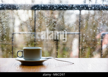 A white espresso cup with a spoon on a wooden table in front of a glass window covered with rain drops with a shallow depth of field. Stock Photo
