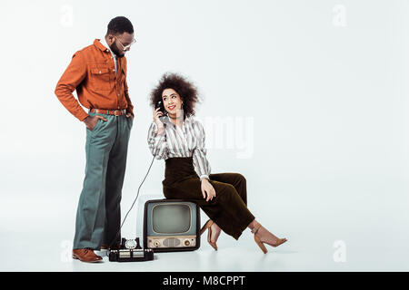 african american retro styled girlfriend sitting on vintage television and and talking by phone on white Stock Photo