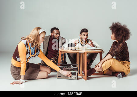 multicultural retro styled friends sitting at table, girl tuning vintage radio Stock Photo