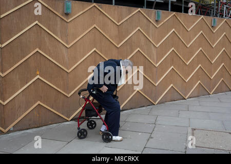 An elderly pensioner struggles to his feet using a four-wheeled rollator mobility aid past the zigzag battens of a construction hoarding at Notting Hill, on 13th March 2018, in London, England. Stock Photo