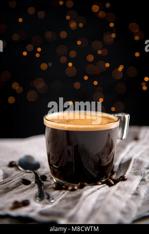 Espresso coffee in a glass cup surrounded by coffee beans and teaspoon on a gray cloth, and blurred lights in the dark background. Stock Photo