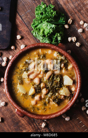 high angle view of an earthenware bowl with kale stew with potatoes and chickpeas, on a rustic wooden table sprinkled with some dry chickpeas Stock Photo