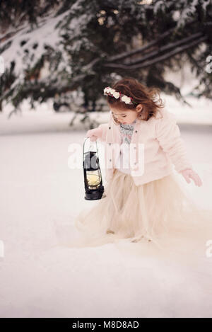 Little girl wearing a beige coat and long dress, looking at lamp with candle standing among the branches, covered with snow in the park in winter time Stock Photo
