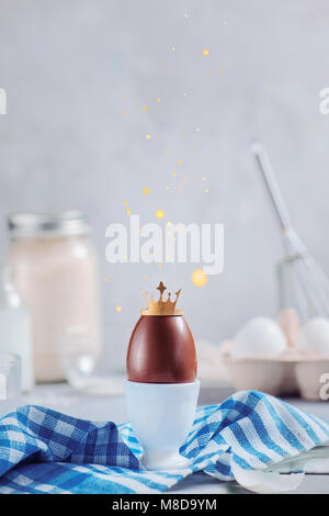 Chocolate Easter egg with golden crown and falling glitter in a porcelain cocotte. High key background with copy space. Royalty and uniqueness concept Stock Photo