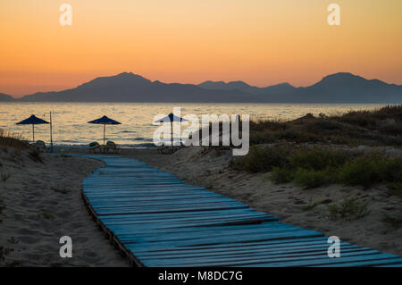A light blue boardwalk leads down to the umbrellas on the beach, as the sun set behind the mountains at golden hour. Stock Photo
