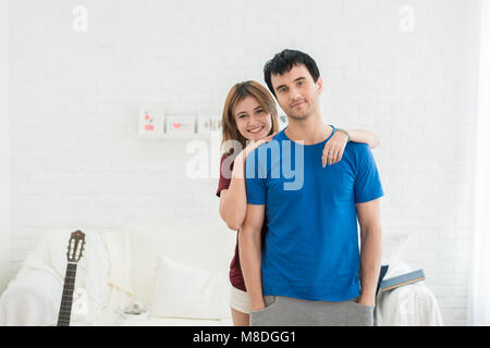 Beautiful young couple is hugging, looking at camera and smiling while standing in living room at home. Couple relax at modern home indoors and have f Stock Photo