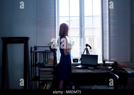 Mid adult woman, standing beside window, using smartphone, camera and laptop on table Stock Photo