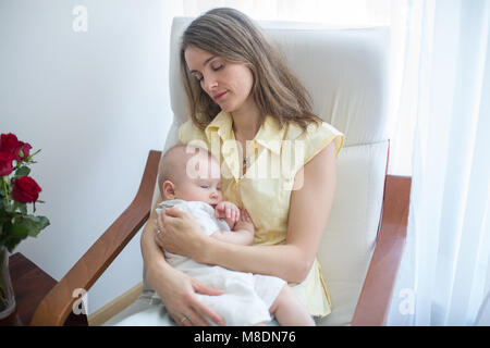 Young mother holding her baby, rocking him to sleep, sitting in armchair in a cozy sunny bedroom Stock Photo