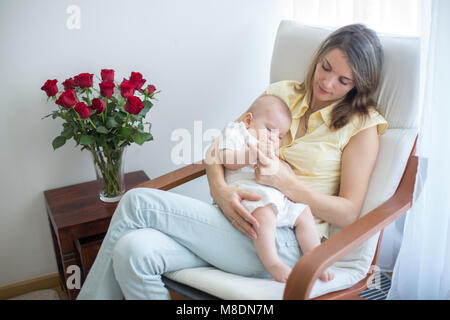 Young mother holding her baby, rocking him to sleep, sitting in armchair in a cozy sunny bedroom Stock Photo