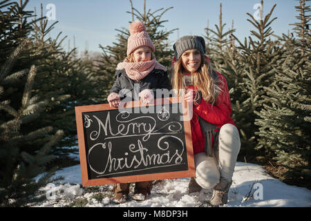 Girl and mother in christmas tree forest with merry christmas sign, portrait Stock Photo