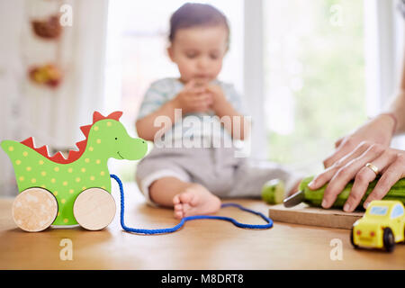 Baby girl sitting on kitchen counter watching mother slicing cucumber Stock Photo