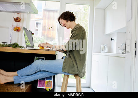 Mid adult woman sitting in kitchen, using laptop, laughing Stock Photo