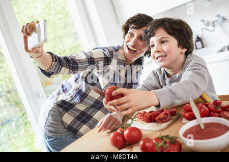 Mother and son preparing food in kitchen, mother taking selfie using smartphone Stock Photo