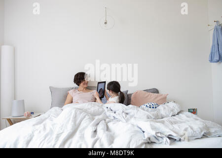 Mother and daughter relaxing in bed, daughter photographing mother using digital tablet Stock Photo
