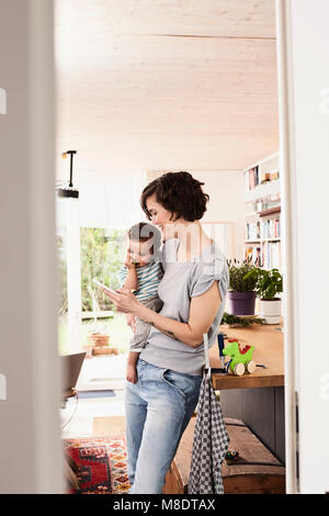 Mother holding baby daughter in kitchen, looking at smartphone Stock Photo