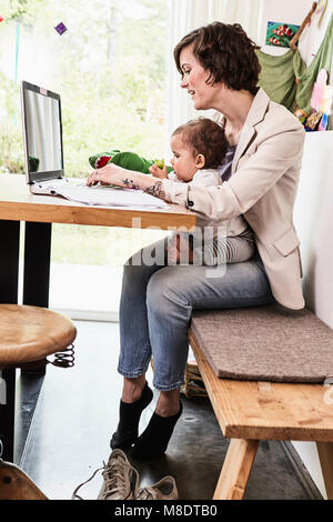 Mother holding sitting with baby daughter, working on laptop Stock Photo