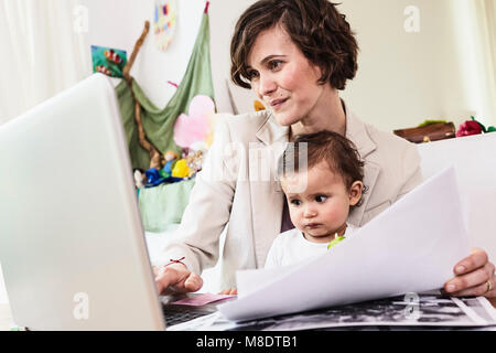 Mother holding sitting with baby daughter, working on laptop Stock Photo