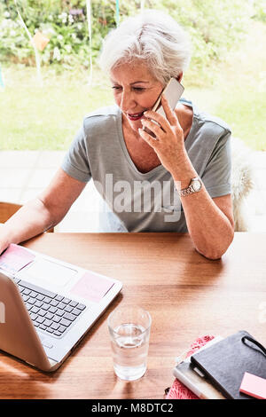 Senior woman sitting at table with laptop in front of her, using smartphone Stock Photo