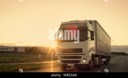 Front-View of Semi-Truck with Cargo Trailer Driving on a Highway. He's Speeding Through Industrial Warehouse Area with Sunset in the Background. Stock Photo
