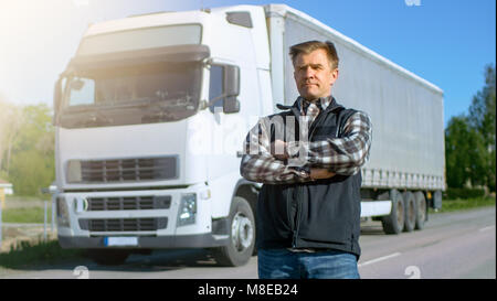 Professional Stands in the Middle of the Road and Proudly Crosses Arms. On the backgroud Parked White Semi Truck with Cargo Trailer Attached. Stock Photo