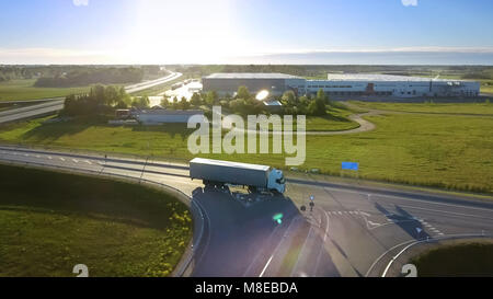 Aerial View of White Semi Truck with Cargo Trailer Passing Highway Overpass/ Bridge. Eighteen Wheeler is New, Loading Warehouses are Seen in the Backg