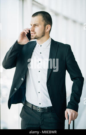 Handsome young man on business trip walking with his luggage and talking on cellphone at airport. Travelling businessman making call. Stock Photo