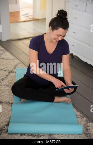 Pregnant woman sitting on yoga mat and using tablet pc Stock Photo