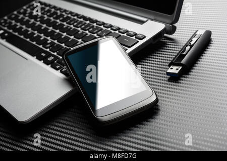 Blank Black Business Smartphone With Blue Reflection Leaning On A Notebook Keyboard Next To An USB Storage Flash Drive, All Above A Carbon Layer Stock Photo