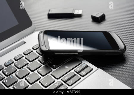 Blank Black Smartphone With Reflection Leaning On A Business Notebook Keyboard Next To An Open USB Storage Flash Drive, All Above A Carbon Layer Stock Photo