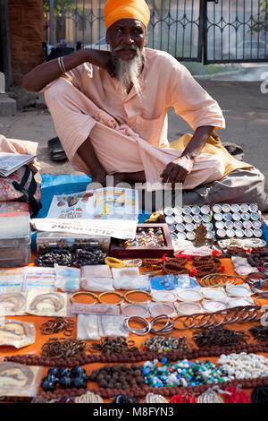 Indian sadhu - holy man sitting on pavement and selling jewelery and mala beads on streets of Jodhpur in India Rajasthan. He is dressed in light pink  Stock Photo