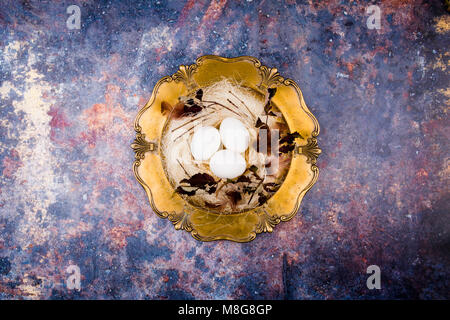 White eggs in stylish straw nest decorated with leaves and feathers on the old golden platter. Old rusty background. Flat lay, top view. Easter concep Stock Photo