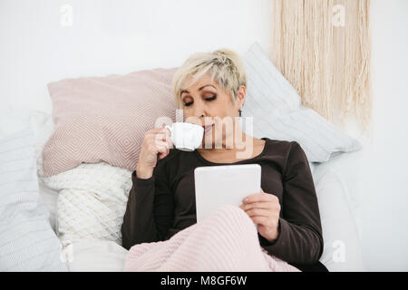 An elderly woman lying in bed drinking morning coffee uses a tablet to view news or chat with friends on a social network. Elderly generation and new technologies. Stock Photo