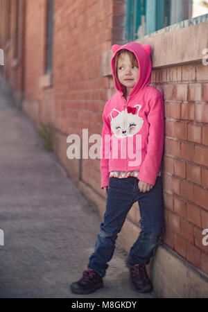 Little red headed girl, leaning against a brick wall, wearing a pink Hello Kitty hoodie.   Canon EOS 6D,  Bausch & Lomb 3 1/4 inch 82mm f/2.8 Petzval  Stock Photo