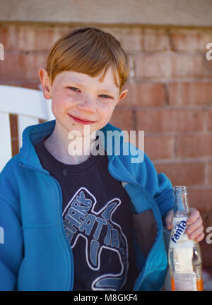 Boy with red hair and freckles, wearing a blue jacket, and holding a bottle of Orange Fanta soda. Stock Photo