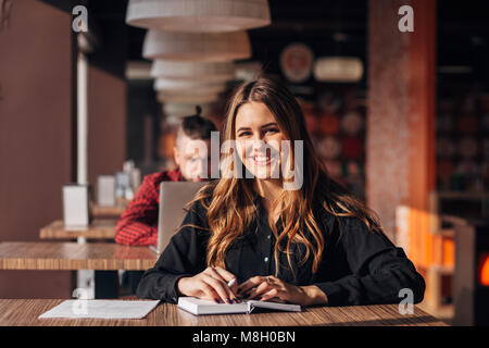 young woman smiling at camera while working remotely in coffee shop. Positive female with notepad Stock Photo
