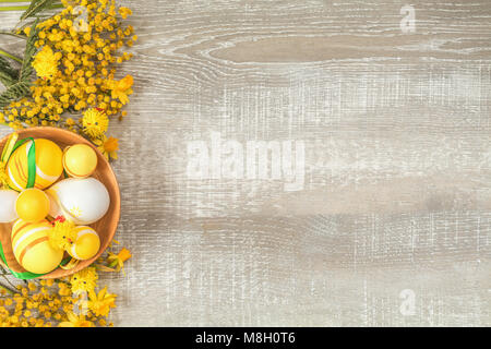 Composition with painted eggs in a wooden bowl. Easter accessories, mimosa and yellow daffodils on a light wooden surface. Yellow orange easter concep Stock Photo