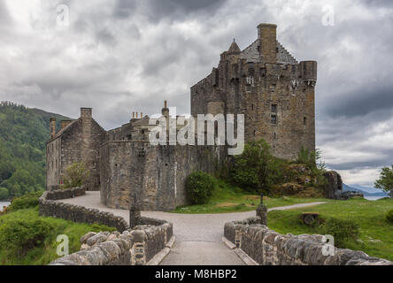 Dornie, Scotland - June 10, 2012: Frontal view of brown stone Eilean Donan Castle, seen from footbridge. Under rain-heavy dark cloudscape. Green veget Stock Photo