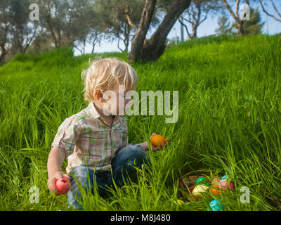 Curious toddler boy having found a basket of colorful Easter eggs in grass Stock Photo