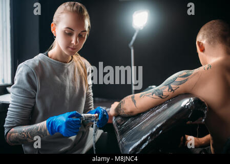 Female tattooist makes tattoo by machine on the shoulder. Professional tattooing in salon Stock Photo