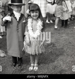 1950s, historical picture of two children standing outside at a fete together taking part in a fancy dress competition...the happy little girl, is proudly wearing her costume of a hula or grass skirt with flower necklace, while the a little boy  isin his costume of a town crier with coat, top hat and bell. Stock Photo