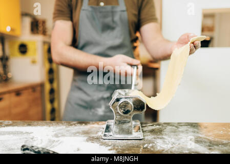Bearded chef in apron works with dough in pasta machine on wooden kitchen table. Homemade spaghetti cooking process Stock Photo