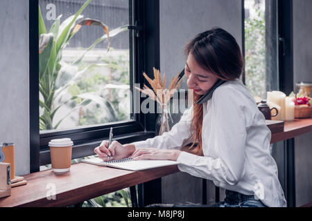 Asian Businesswoman use mobile phone contact customer and writing a note on wood table at coffee shop near window,woking outside office lifestyle Stock Photo