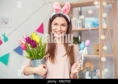 Young woman at home easter celbration concept wearing bunny ears holding decorative eggs on sticks and bouquet of tulips looking camera smiling cheerf Stock Photo