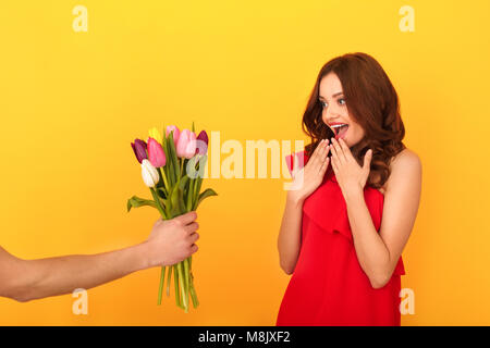 Young woman studio isolated on yellow wall wearing red dress getting tulips bouquet from handsome man surprised happy Stock Photo