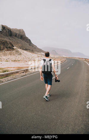 Traveler walks in the center of an epic winding road. Huge volcanic mountains in the distance behind him. Sao Vicente Cape Verde Stock Photo