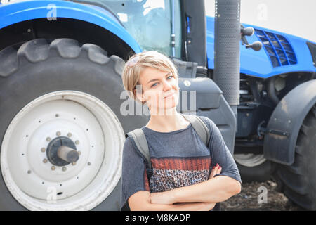 Pretty young woman standing in field with tractor in background Stock Photo