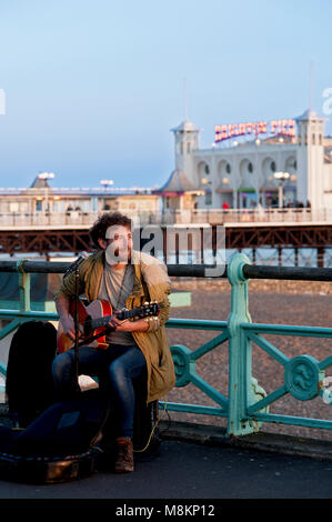 A busker entertains passersby with songs on Brighton's promenade near the famous Brighton Pier, one of the UK's most popular tourist destinations. Stock Photo
